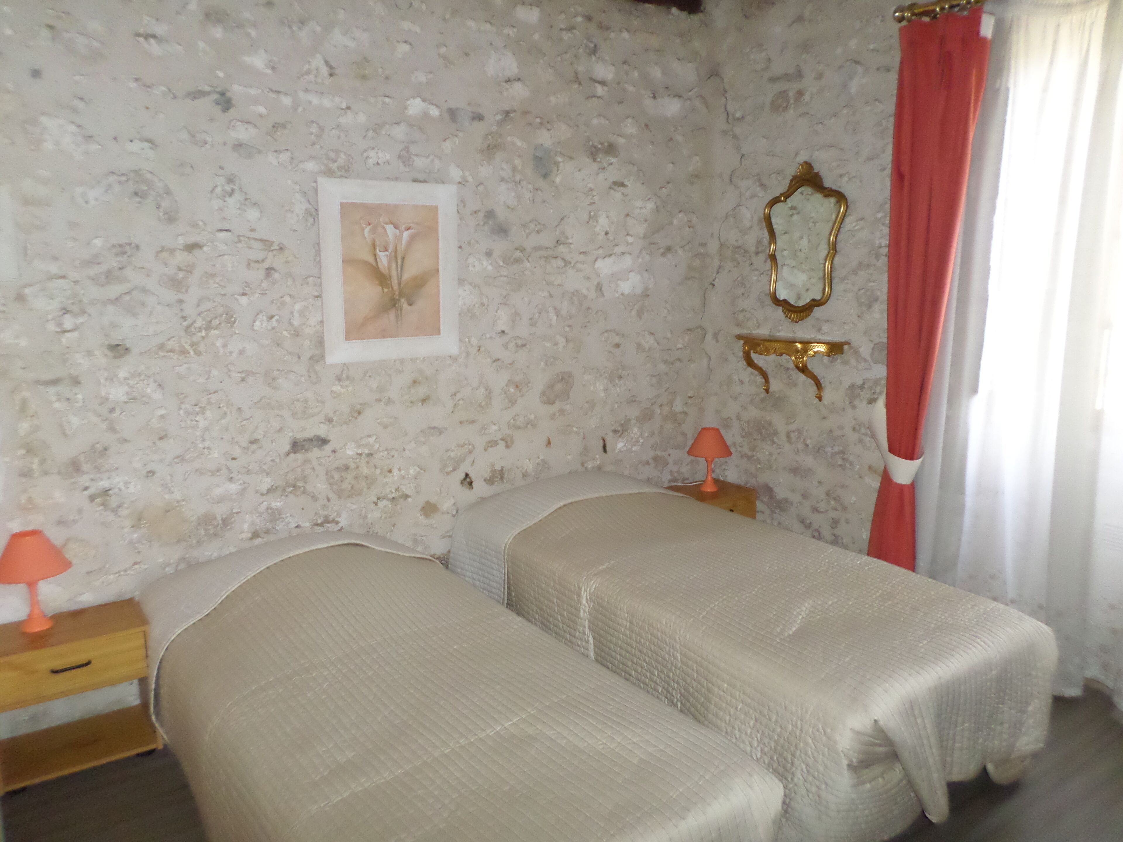 A room with two beds, a wooden nightstand with a lamp, a stone wall, a framed picture, and a mirror.