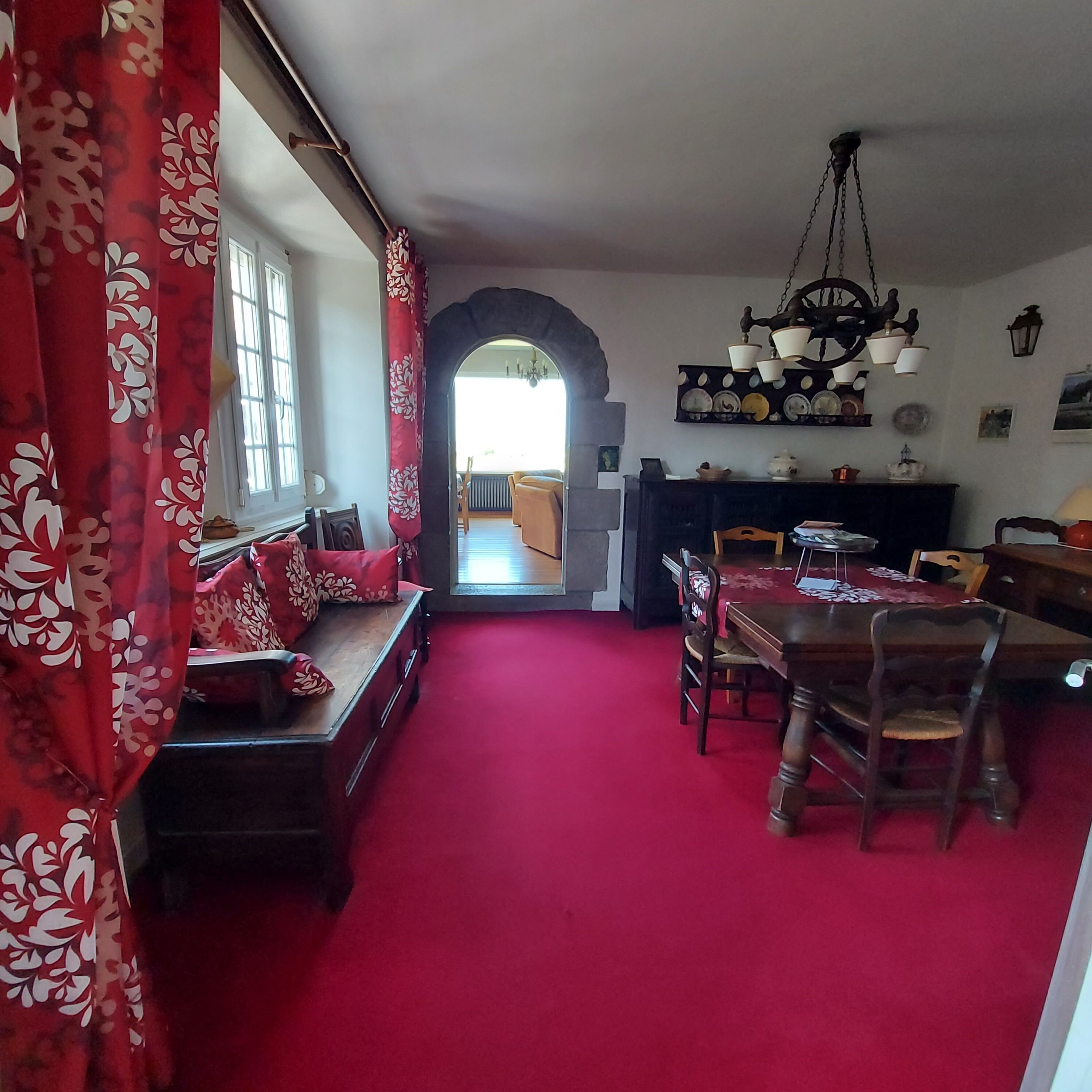 Een eetruimte met een rood tapijt, houten meubels en een doorkijk naar een andere kamer via een boog.