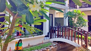 Un jardin avec un pont de pierre, une végétation luxuriante et un bâtiment doté de balcons.
