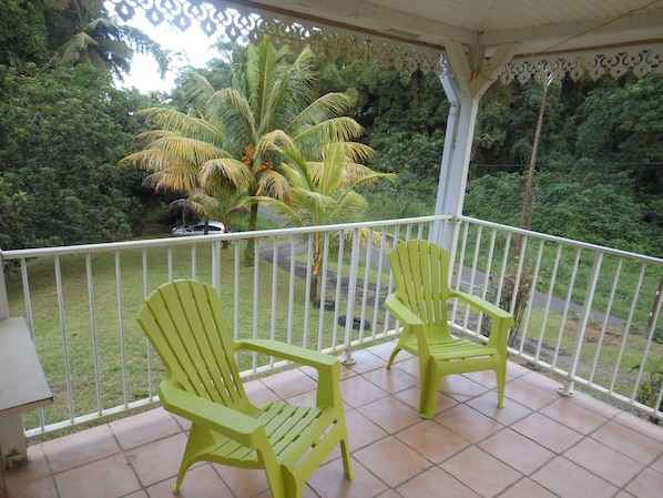 A covered balcony with two green chairs, a white railing, and a view of a road and greenery. - Villa F3 in SAINTE-MARIE (SAINTE-MARIE)