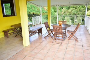 A covered patio with a table and chairs, benches, and a window overlooking greenery.