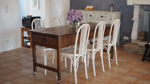 Uma área de refeições com mesa de madeira e cadeiras brancas, um vaso com flores e um armário branco com um quadro na parede.
