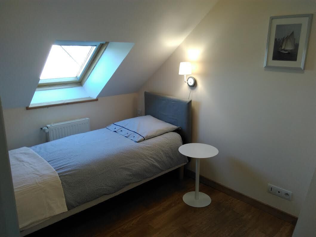 A small, single-bed room with a skylight, a bedside table, and a wall-mounted lamp.