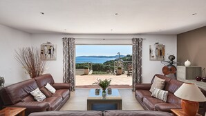Een ruime woonkamer met een groot raam dat uitzicht biedt op zee en heuvels. Er zijn twee leren banken, een salontafel en een lamp.