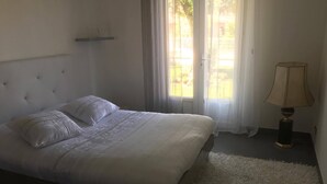 Un dormitorio con cama, lámpara de noche, ventana con cortinas y vistas al exterior.