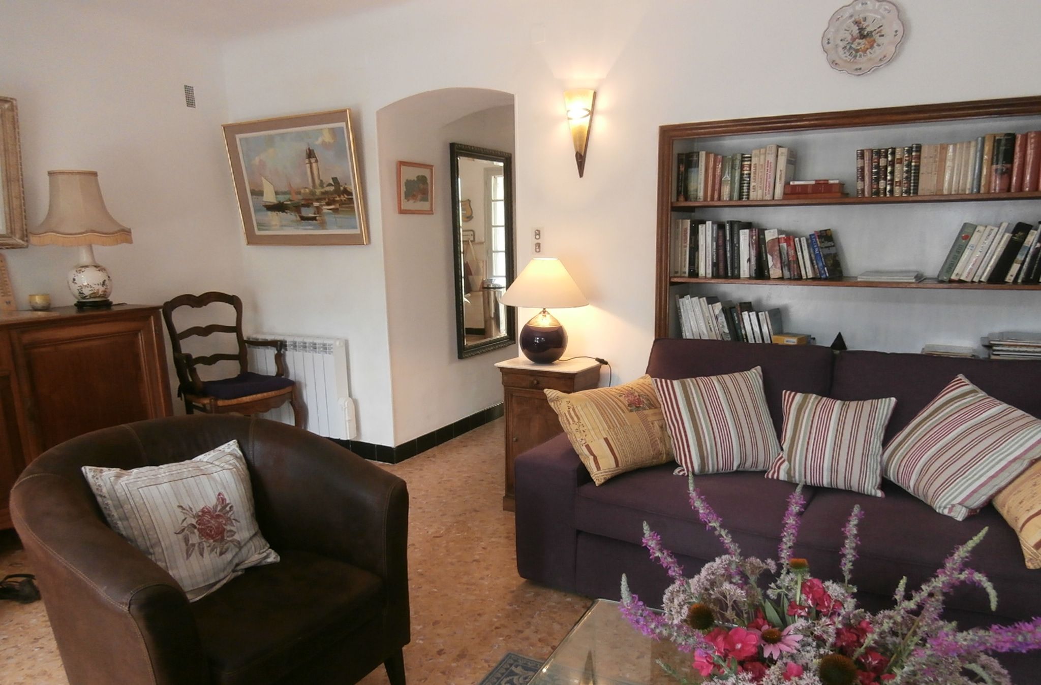 A living room with a brown leather armchair, a purple sofa, a glass coffee table, a bookshelf, a lamp, and a painting on the wall.
