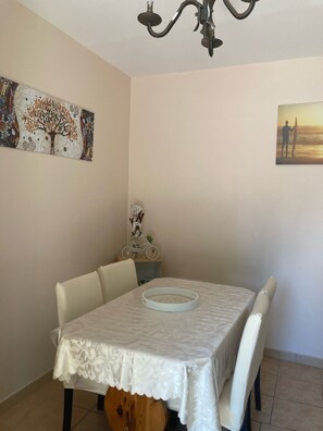 Un comedor con una mesa cubierta por un mantel blanco, rodeada de sillas. Un candelabro cuelga sobre la mesa, y se exhiben obras de arte en las paredes.
