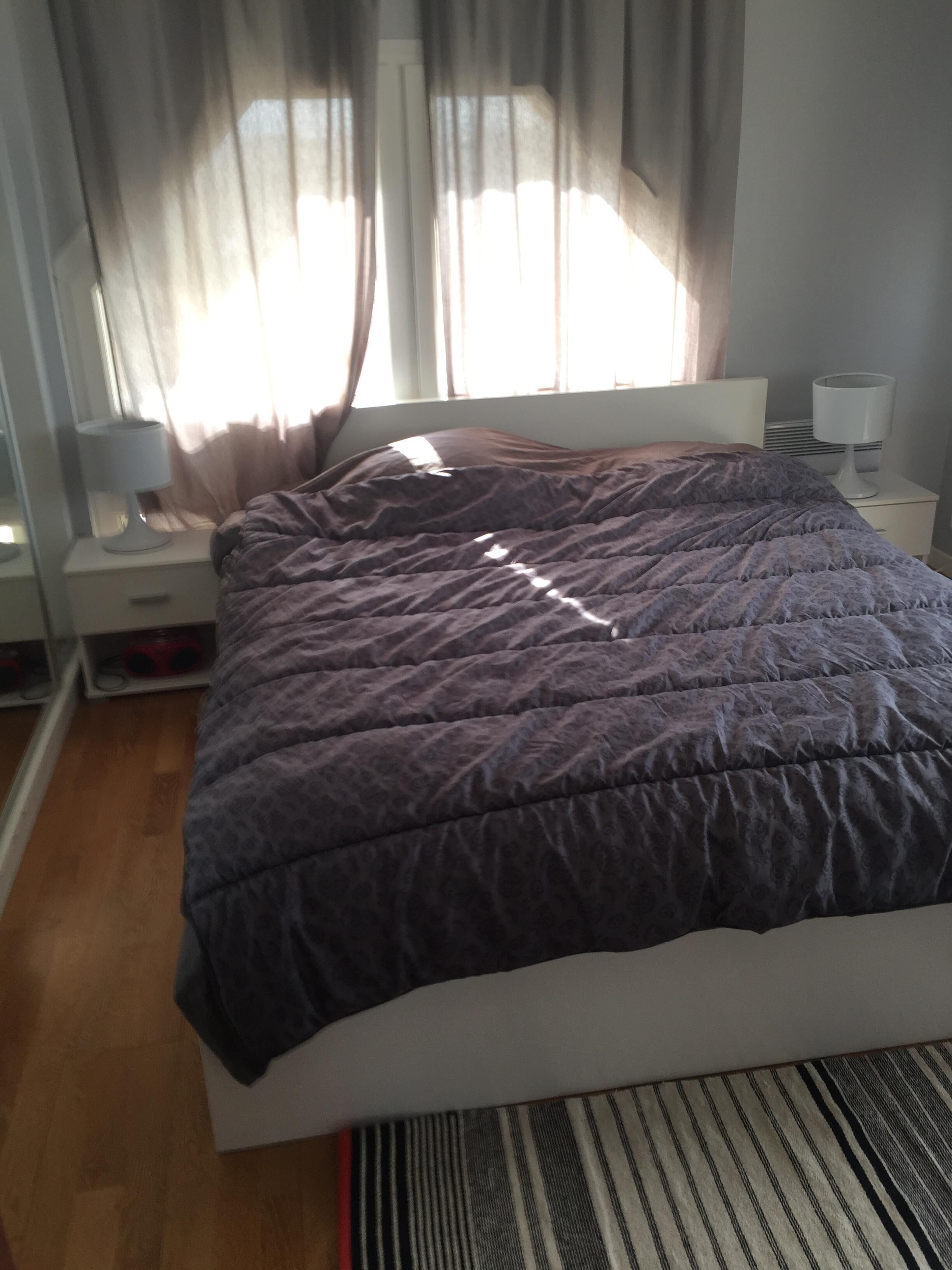 Un dormitori amb un llit, una tauleta de nit, una làmpada i una finestra amb cortines.