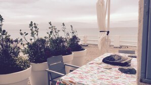 Terraço à beira-mar com plantas em vasos, uma mesa com toalha de peixe e um guarda-sol branco.