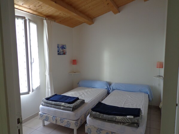 A room with two beds, a window with curtains, a small table with a lamp, and a painting on the wall.