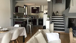 Una cocina moderna con una isla central, un comedor con mesa y sillas, y una escalera que sube a otro nivel.