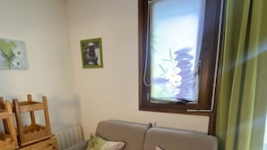 A cozy living room with a grey sofa, wooden side tables, and a window with a view of flowers. - COQUET STUDIO FEET OF THE TRACKS WELL EQUIPPED EXCEPTIONAL !!! ! (Risoul)