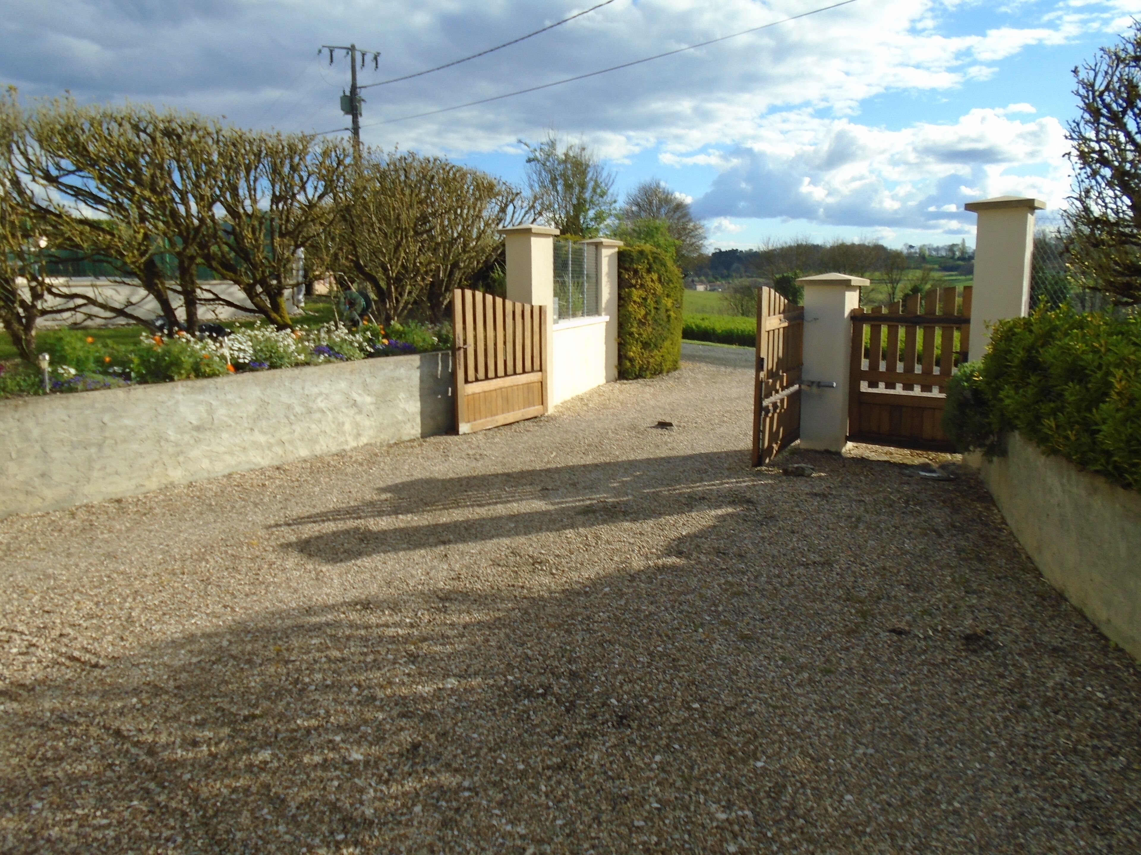 Un camino de grava que conduce a una entrada porticada, con valla de madera y muro de concreto.