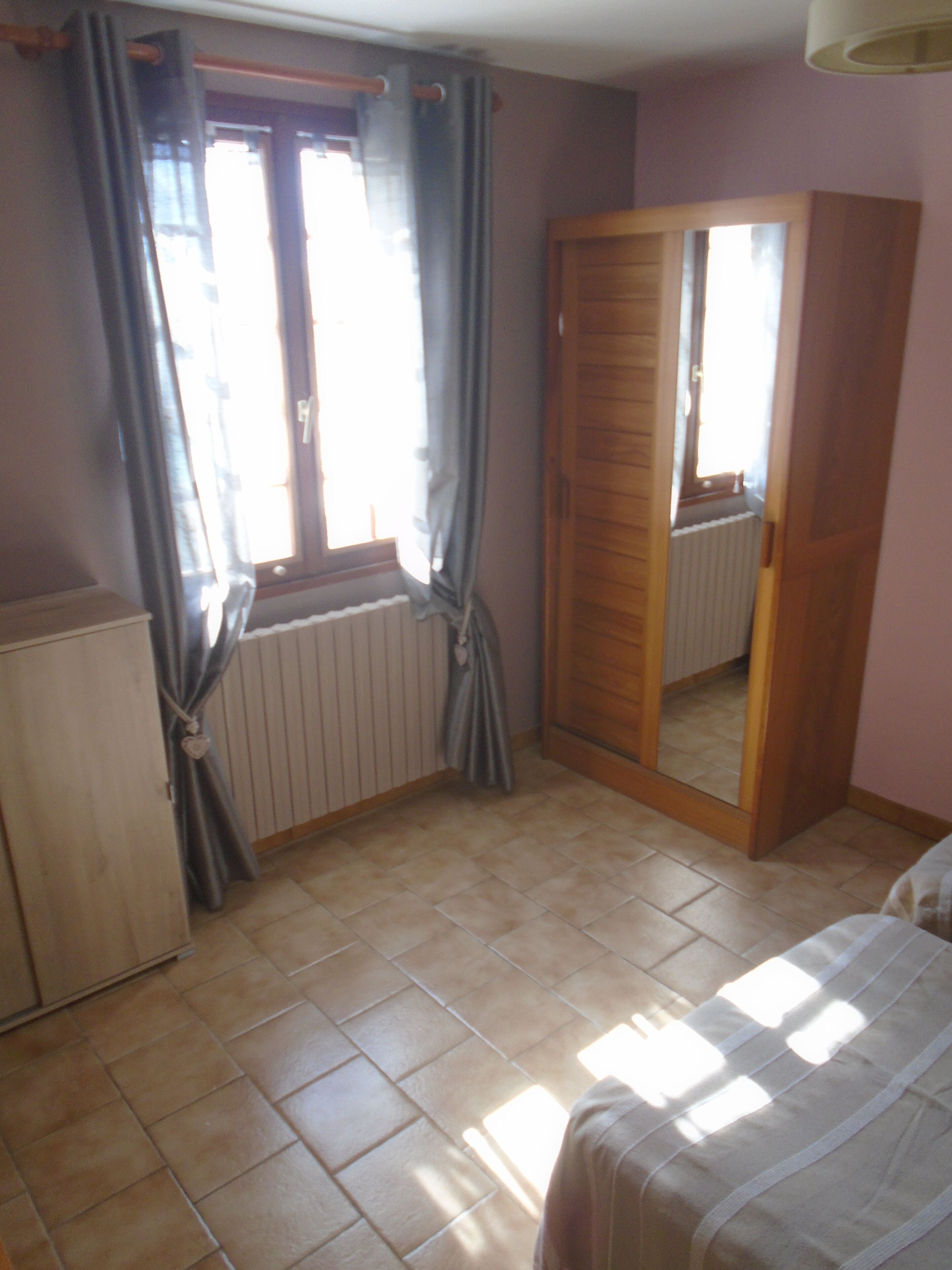 Ein Schlafzimmer mit Bett, Schrank, Heizkörper und einem Fenster mit Vorhängen.