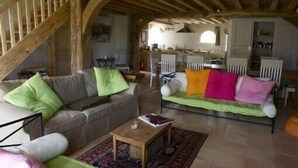 Un salon rustique avec un escalier en bois, un canapé agrémenté de coussins colorés, une petite table en bois et, au fond, un coin cuisine.