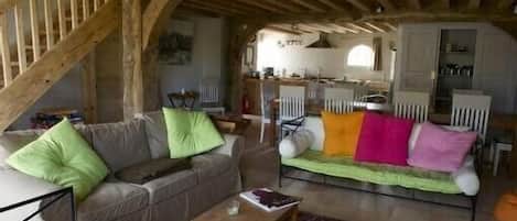 A rustic living room with a wooden staircase, a sofa with colorful cushions, a small wooden table, and a kitchen area in the background.