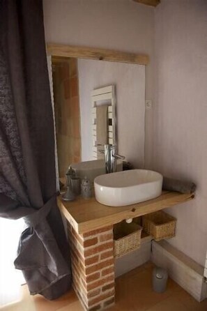 Een badkamer met een wit wastafel, een spiegel en een houten plank. Daaronder staan rieten manden en in de hoek staat een prullenbak.