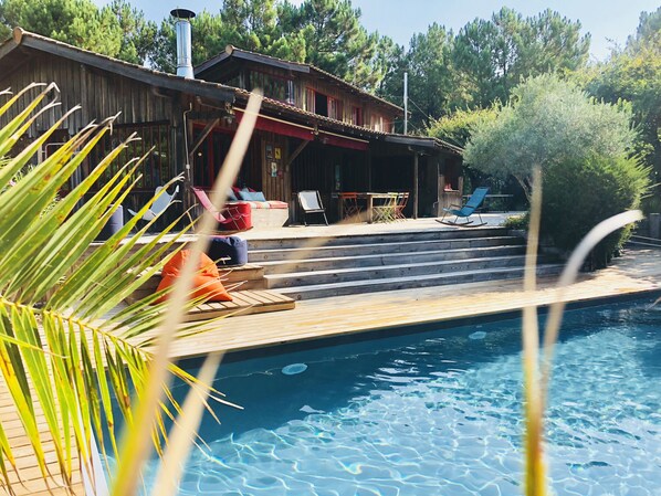 A wooden deck with a pool, outdoor seating, and a building with a red awning. - Large charming cabin (Lège-Cap-Ferret)