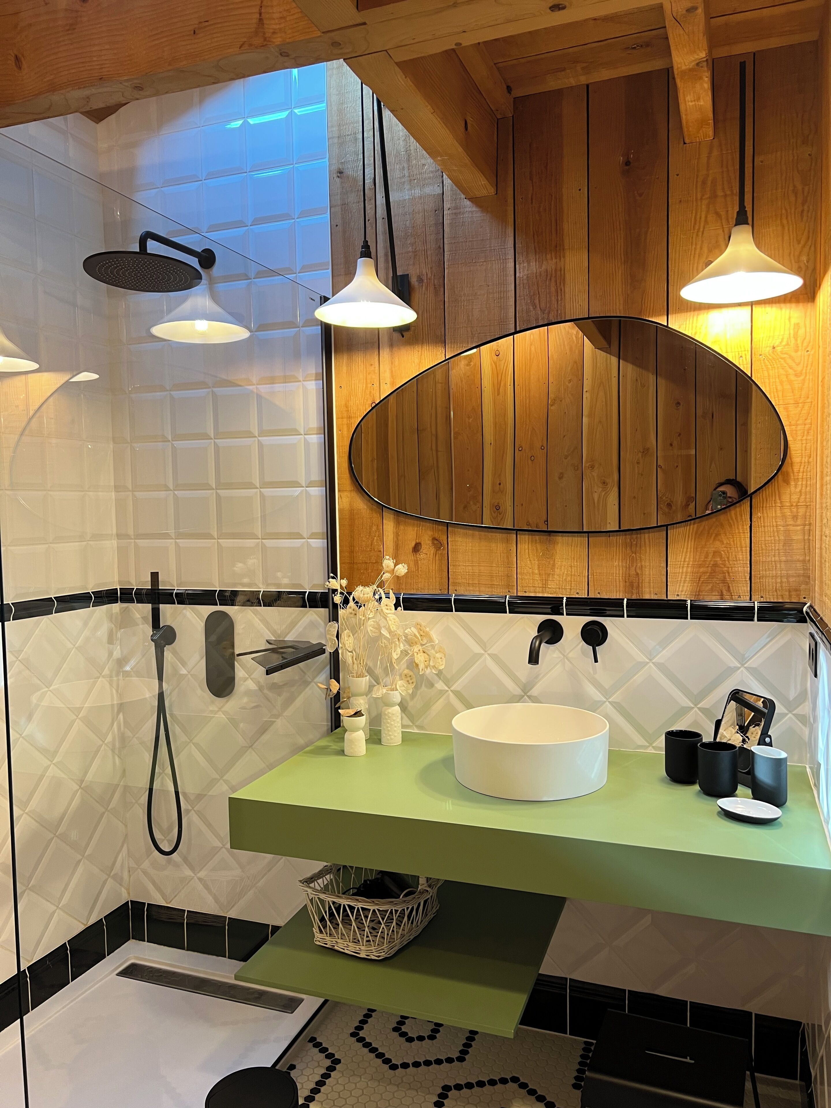 A bathroom with a green countertop, a white sink, and a large mirror. There are three pendant lights, a showerhead, and a towel rack.