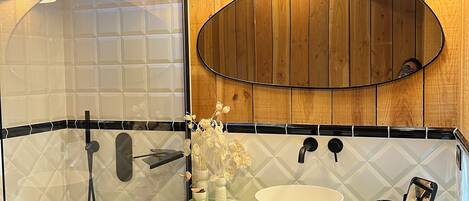 A bathroom with a green countertop, a white sink, and a large mirror. There are three pendant lights, a showerhead, and a towel rack.