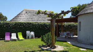 Een achtertuin met een pergola, een kleine boom en een gebouw met rieten dak. Onder de pergola staan plastic stoelen en een tafel.