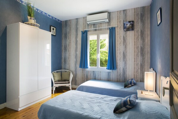Ein Schlafzimmer mit zwei Betten, einem weißen Kleiderschrank, einem Fenster mit Vorhängen, einem Sessel und einer Nachttischlampe.