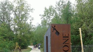 Een metalen bord met de tekst 'PARC AUSONE', een verhard pad en een hek van gaas met bomen op de achtergrond.