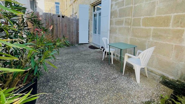 A courtyard with a gravel ground, a wooden fence, and a white door. There are two white plastic chairs and a small table.