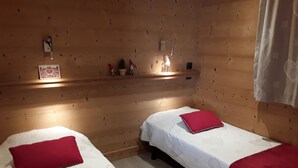 A cozy room with two single beds, each with a red pillow and white bedding. There is a shelf with decorative items and a small window with curtains. - Apartment in the owner's chalet in CREST-VOLAND (COHENNOZ)