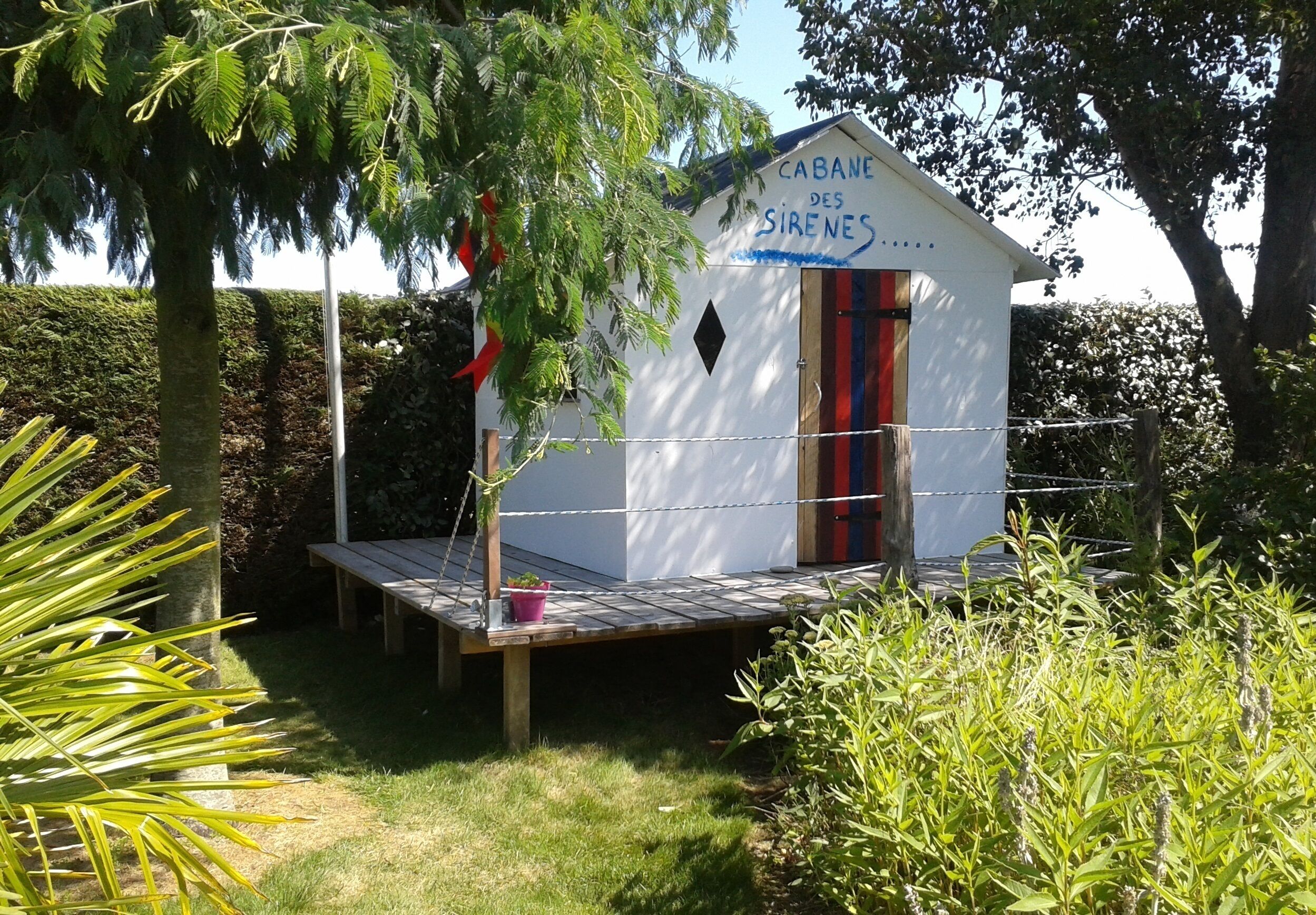 A small, white, single-room cabin with a red and blue striped door, labeled "CABANE DES SIRENES," surrounded by lush greenery.