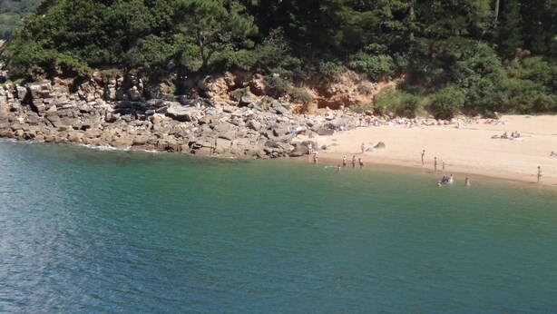 A beach with clear turquoise water, rocky shoreline, and a sandy area with people.