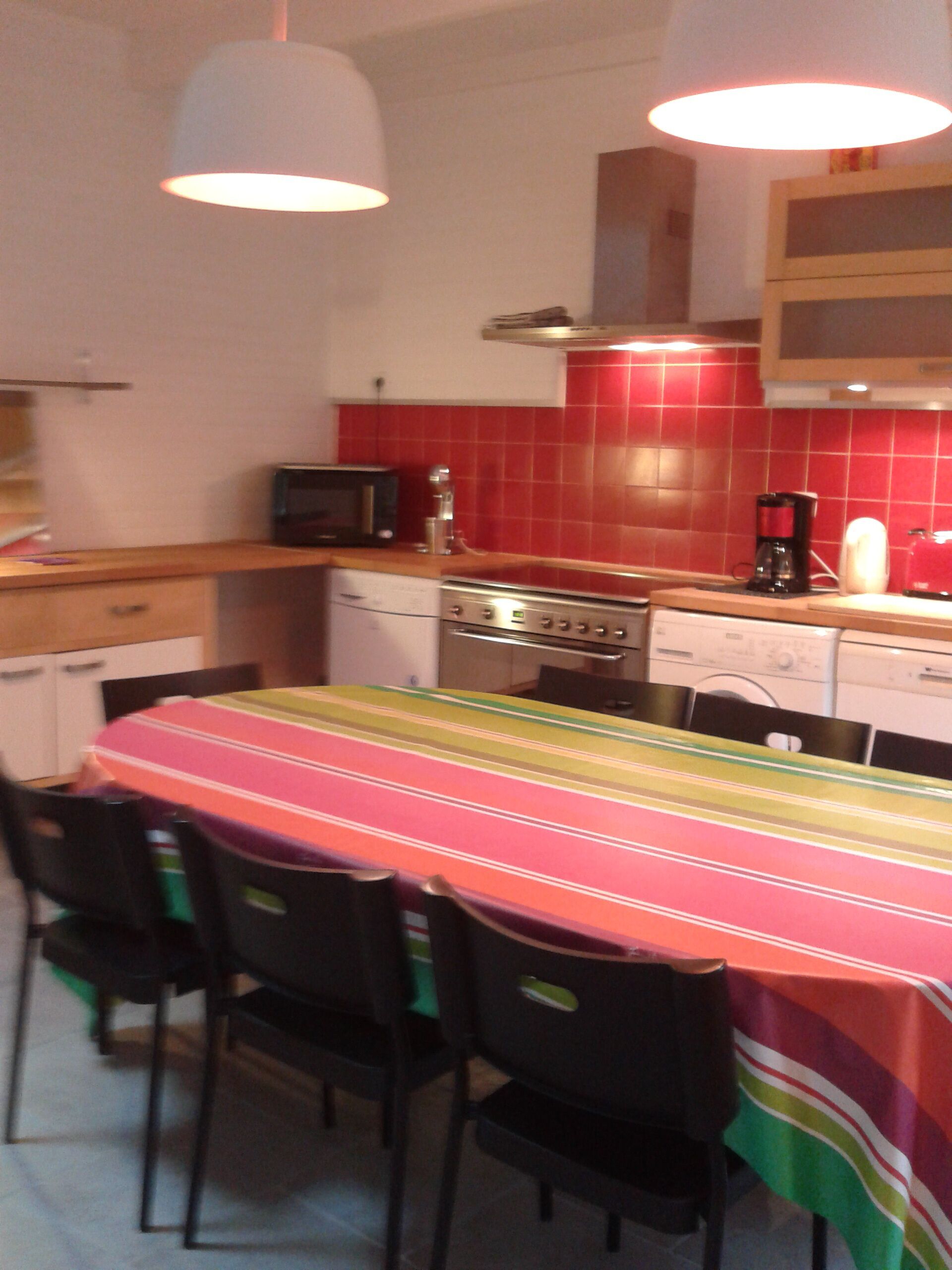 A kitchen with red tiles, a microwave, a coffee maker, and a stove. There is a table with a colorful tablecloth and four chairs.