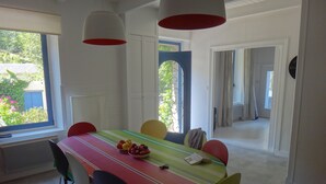 A dining area with a long table, colorful chairs, and a fruit bowl. There are two hanging lights, a window with a view of greenery, and a door leading to another room.
