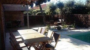 Une terrasse extérieure avec une piscine, des meubles en bois et un espace jardin agrémenté de plantes et d’un arbre.