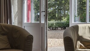 Una stanza con due poltrone beige, una porta bianca e vista su un vialetto sterrato e vegetazione all'esterno.