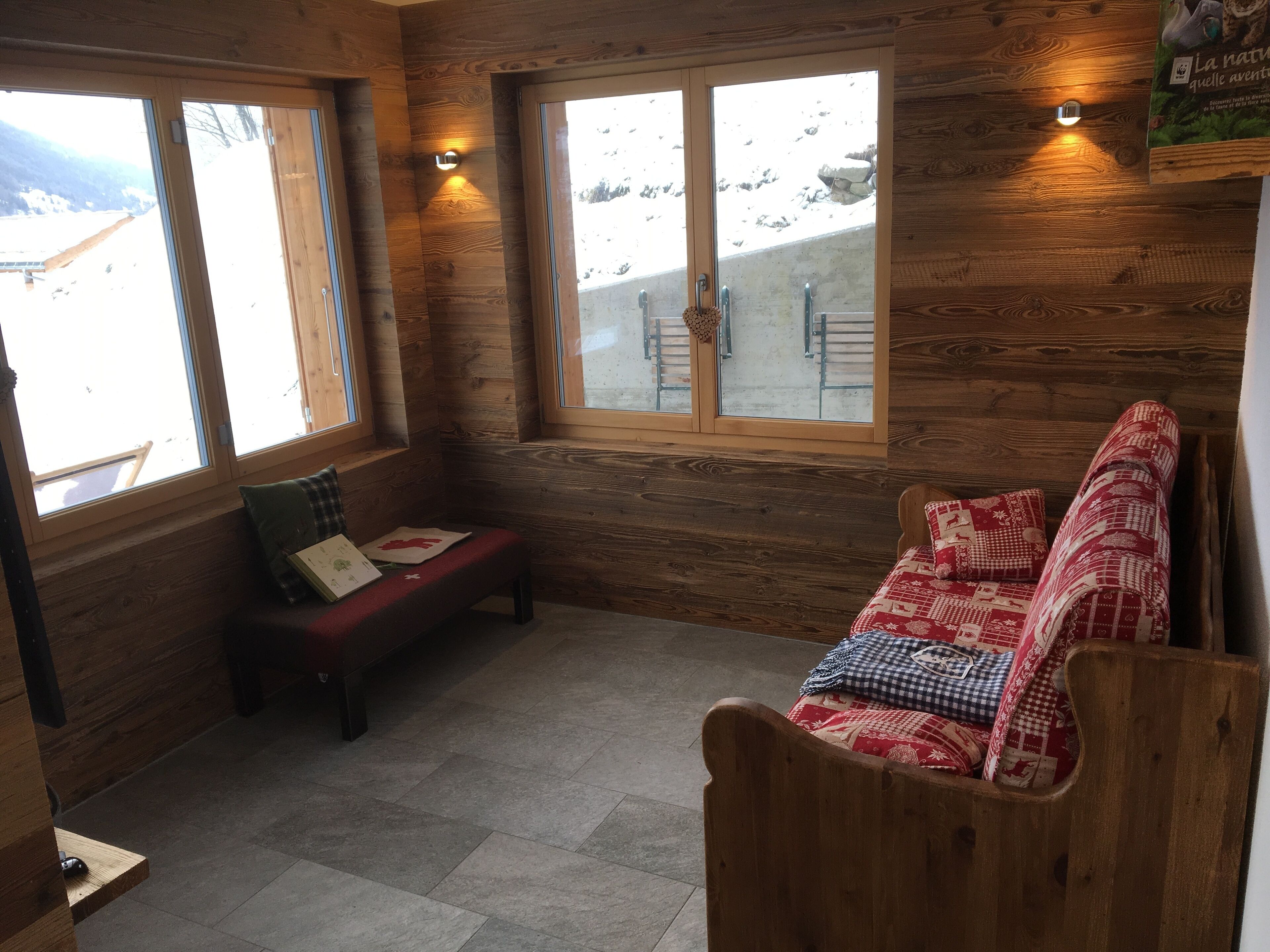 Eine Holzhütte mit einer Bank und einem Sofa, große Fenster mit Blick auf Schnee und eine Holzbalkendecke.