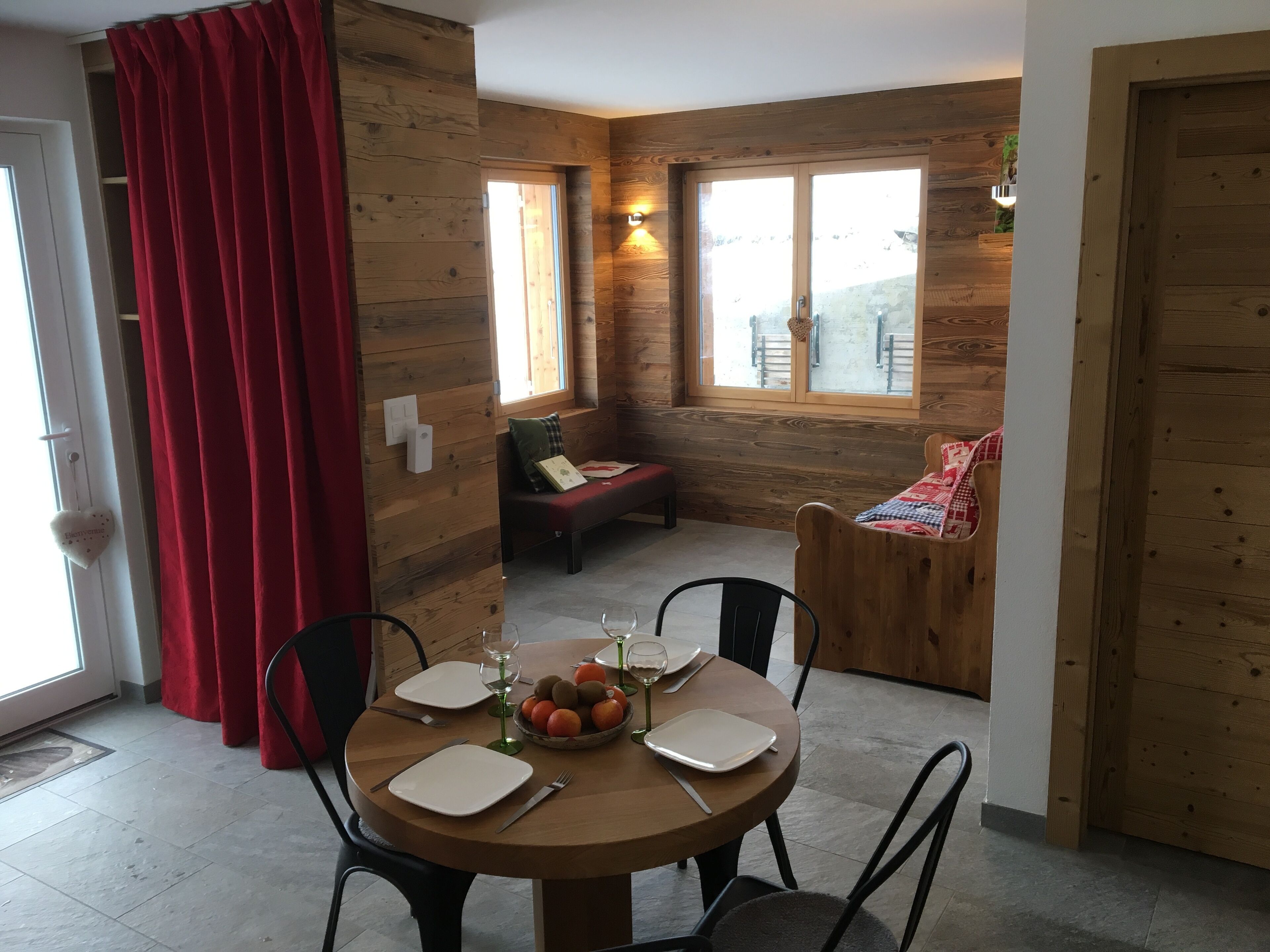 A cozy dining area with a wooden table set for four, surrounded by chairs. A bench with a red cushion is visible in the background, and a window provides natural light.