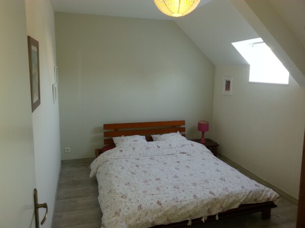 Ein Schlafzimmer mit schrägen Wänden, einem hölzernen Bettgestell, einer blumigen Bettdecke, einer Nachttischlampe und einem kleinen Fenster.
