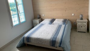 A bedroom with a wooden sloped ceiling, a bed with patterned bedding, two bedside tables with lamps, and a window with blue curtains. - La Fleur de Sel (La Flotte)