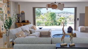 A modern living room with a large sofa, a coffee table, and a TV. There's a balcony with a view of the city and hills. - Luxury single-storey Villa on French Riviera , View & heated swimming pool (ST PAUL DE VENCE)