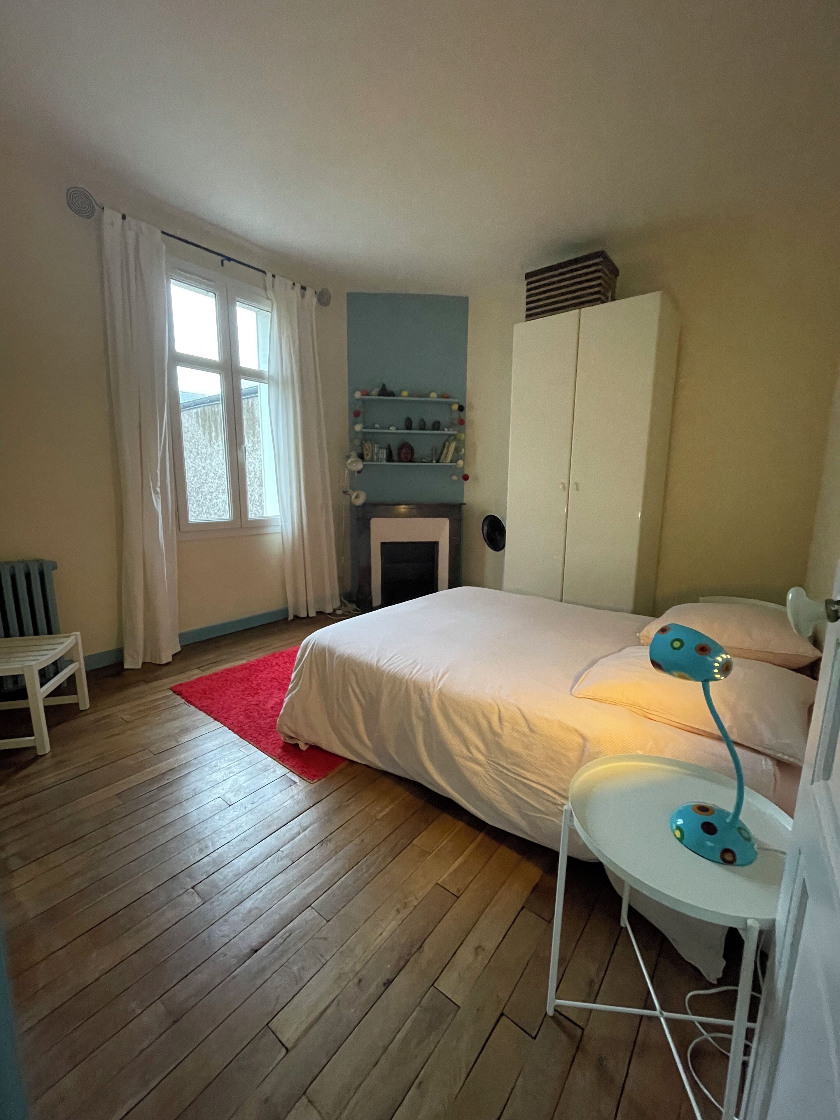 A bedroom with a bed, a red rug, a white chair, a desk with a lamp, a wardrobe, and a window with curtains.