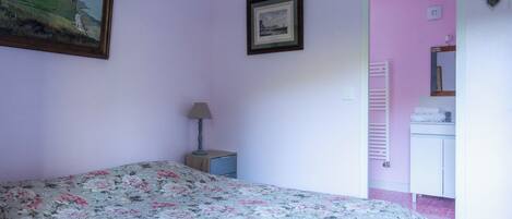 A bedroom with a floral bedspread, a nightstand with a lamp, a framed picture on the wall, and a pink wall.