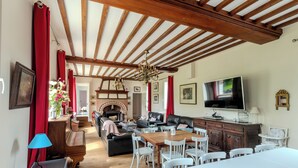 A spacious living room with a wooden ceiling, a fireplace, a TV, and a dining area with white chairs and a table. - Villa Mathilde, a 2-minute walk from the beach (Saint-Pierre-en-Port)