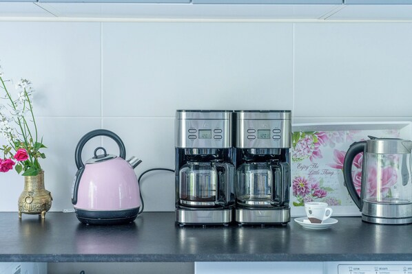 A kitchen counter with a pink kettle, two coffee makers, a floral mug, and a brass vase with flowers.