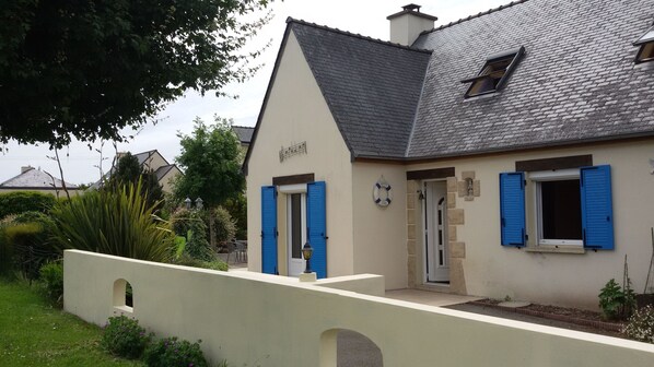A house with a sloped roof, blue shutters, and a small garden area with a pathway leading to the entrance.