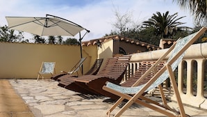 Een terras met stenen vloer, houten lounge stoelen en een witte parasol.