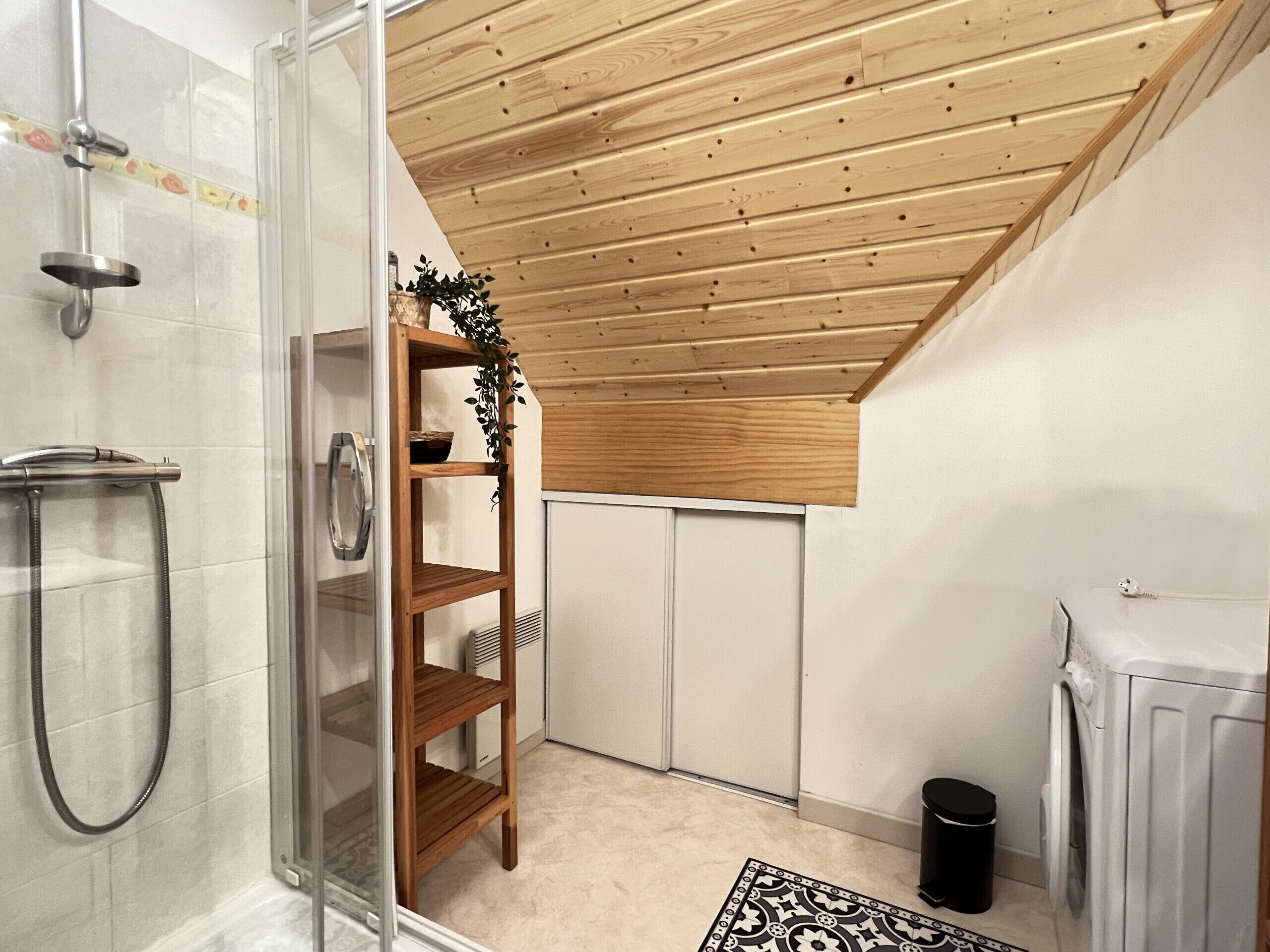 A modern bathroom with a glass shower enclosure, wooden shelving, a radiator, and a washing machine.