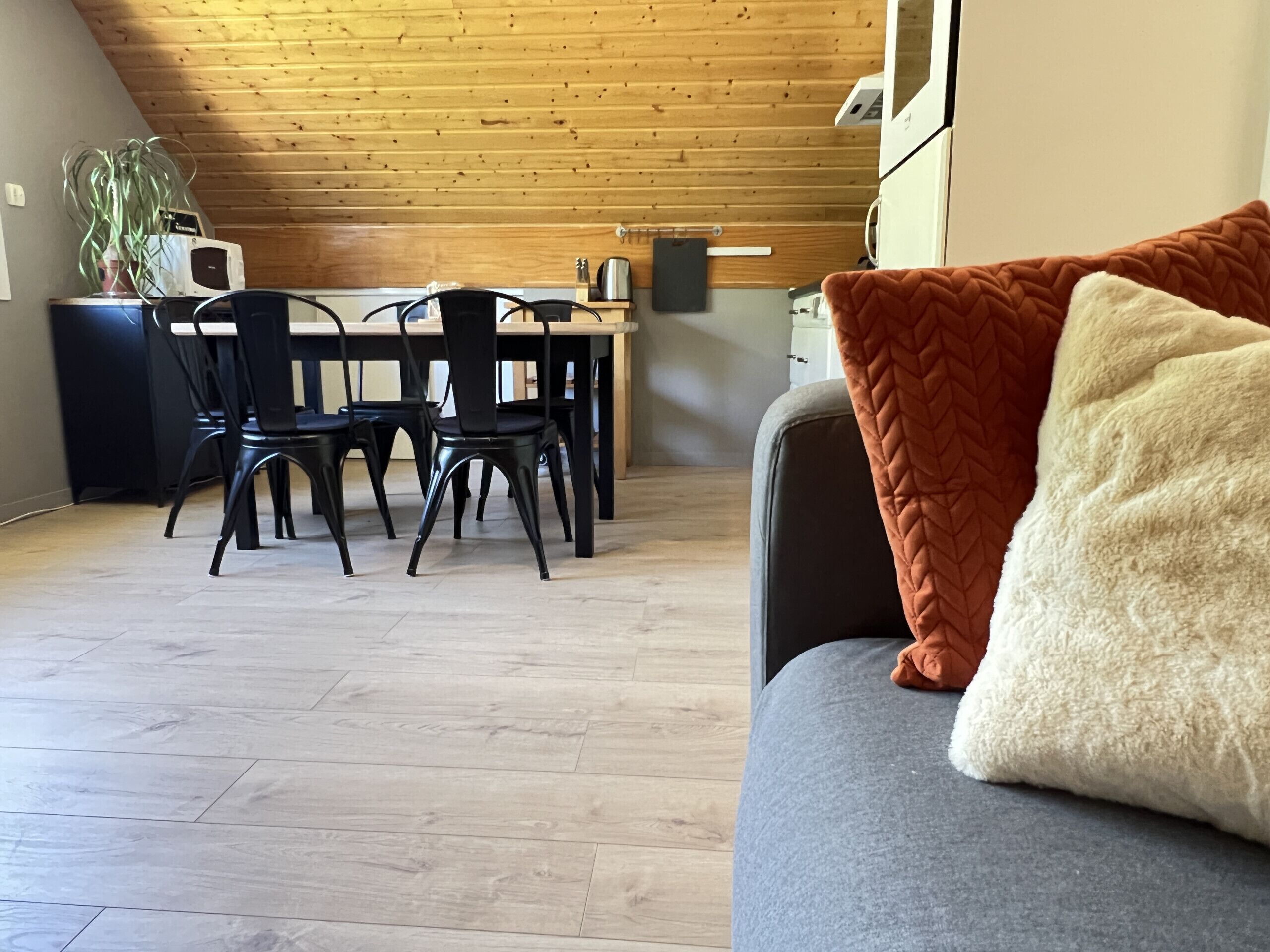 A modern kitchen with wooden ceiling, a dining area with black chairs, and a grey sofa with a white and orange pillow.