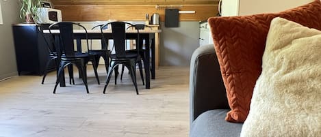 Une cuisine moderne avec un plafond en bois, un coin repas avec des chaises noires et un canapé gris agrémenté d’un coussin blanc et orange.