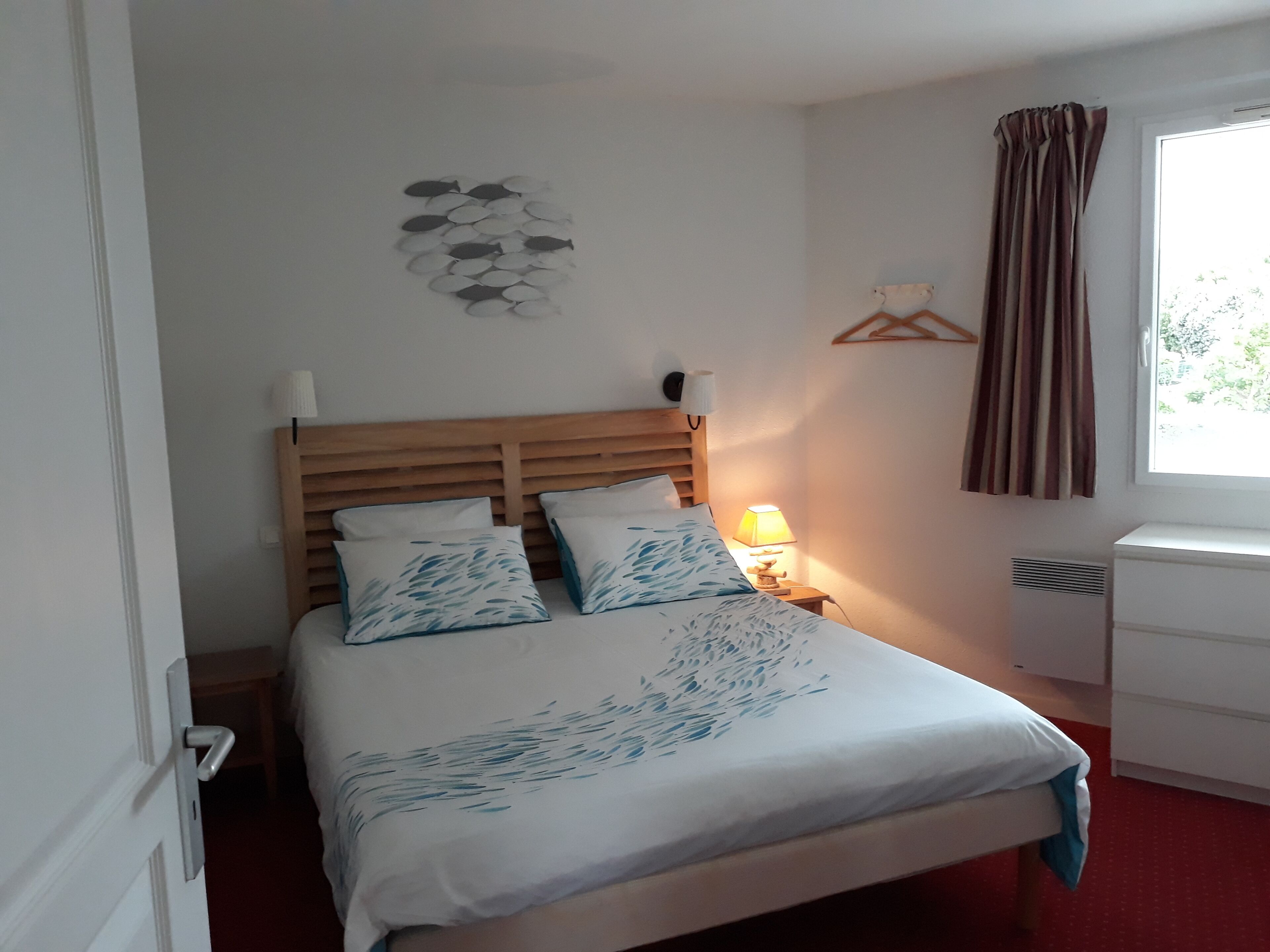 A neatly made bed with a wooden headboard, a bedside lamp, and a wall-mounted shelf with a hanging garment.